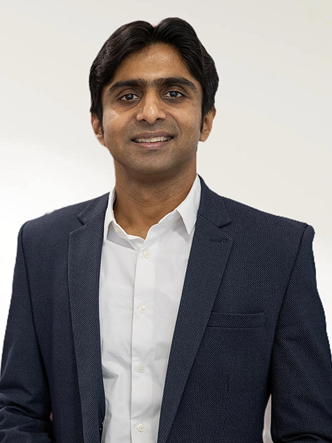 Portrait of a man wearing a navy suit jacket and white shirt, smiling against a light neutral background.